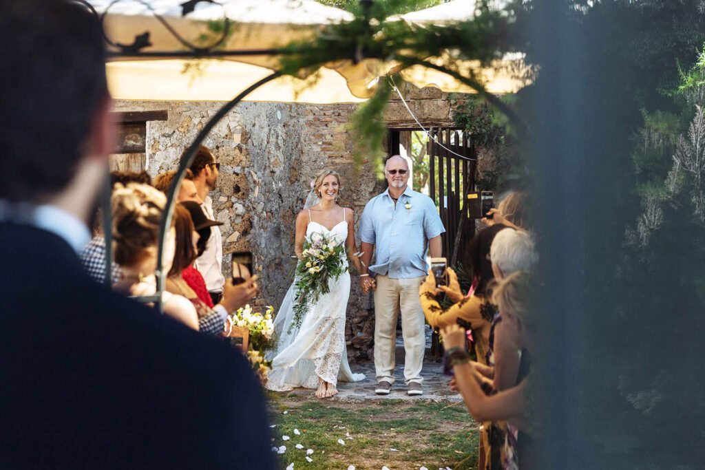 Bride Amy walking down the aisle with her father during the outdoor ceremony at Borgo di Tragliata.