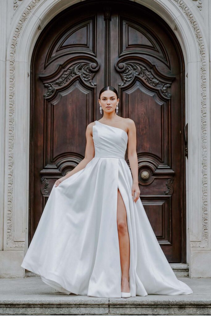 Editorial bridal portrait of elegant bride in one-shoulder luxury wedding dress standing in front of ornate Italian wooden doors