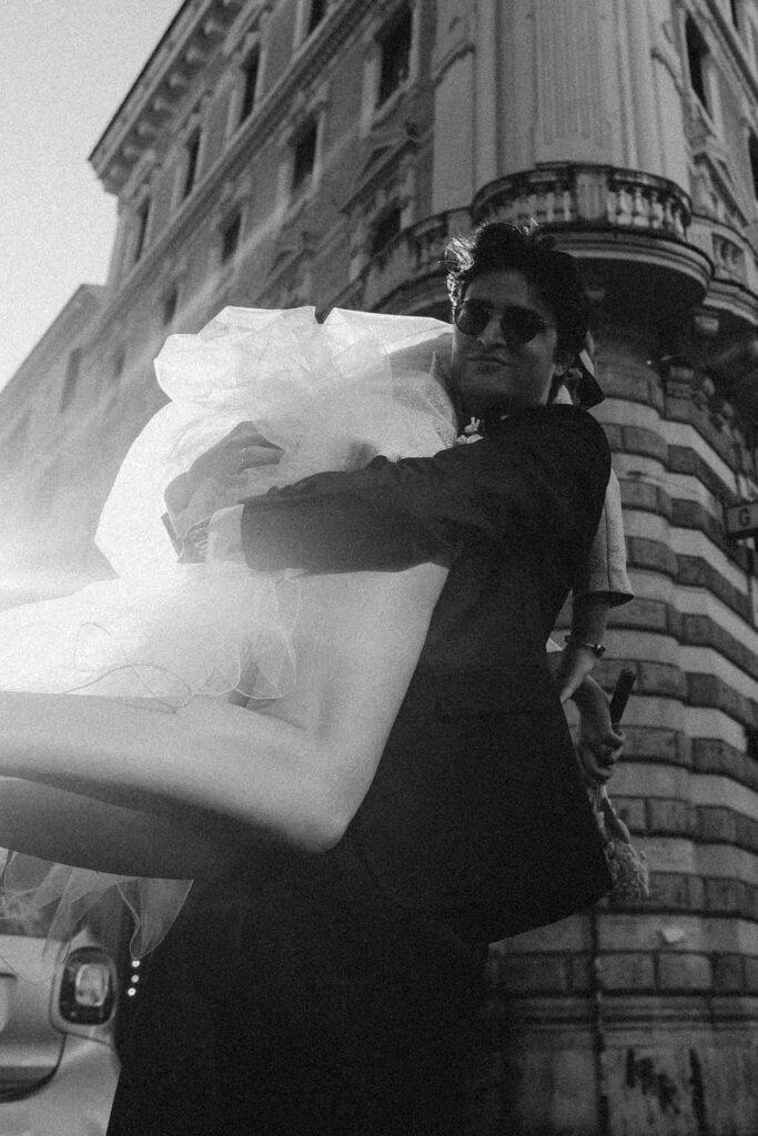 Black and white editorial wedding photo in Rome of groom carrying bride in front of historic Italian architecture