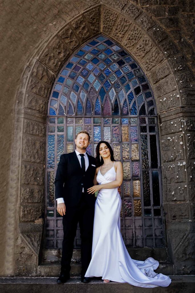 Elegant wedding couple posing under historic stone arch with stained glass window during European destination wedding