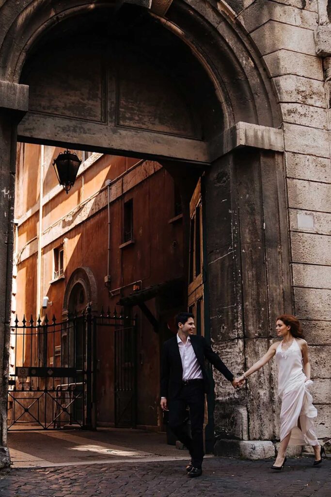 Elegant couple walking hand in hand beneath a historic stone archway in Rome during a destination wedding
