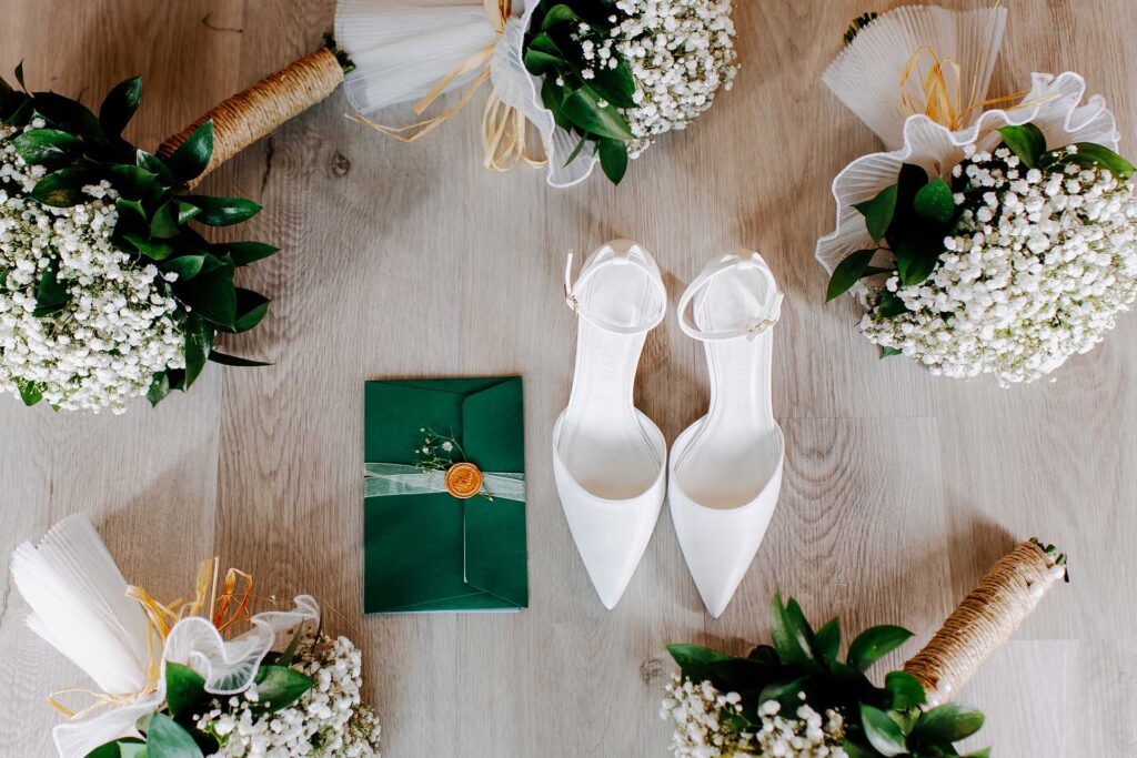 Luxury wedding flat lay featuring white bridal shoes, green envelope invitation and floral arrangements