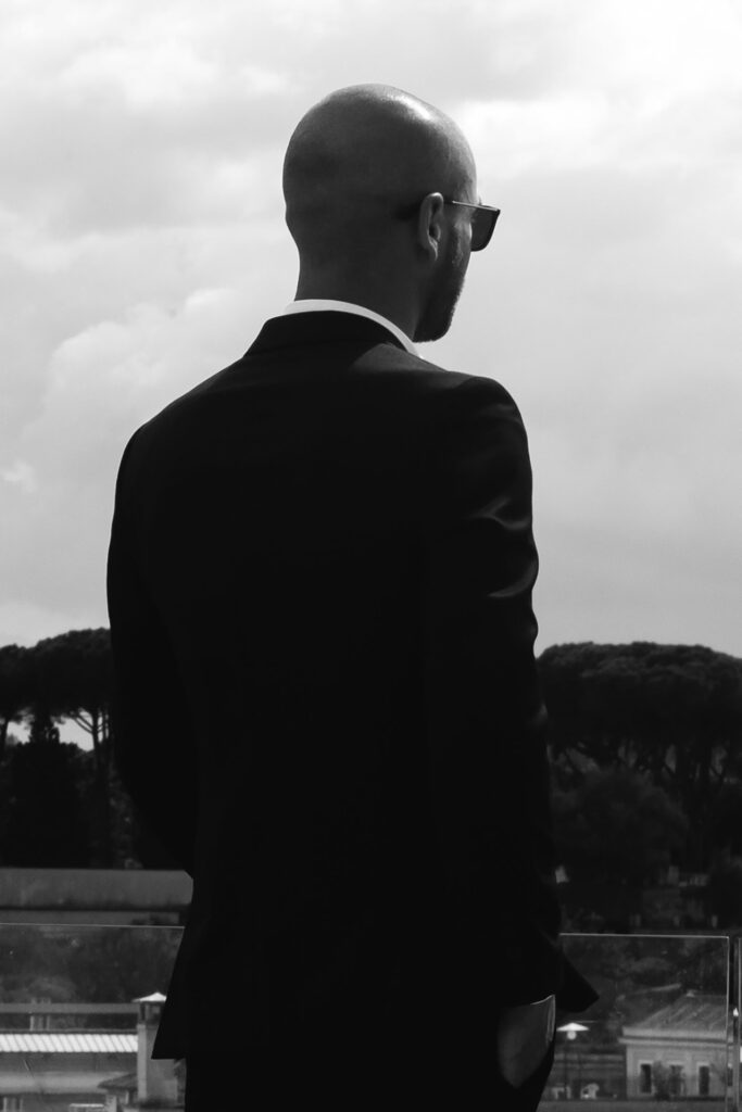 Black and white portrait of a groom in a black suit overlooking Rome from Les Etoiles Terrace