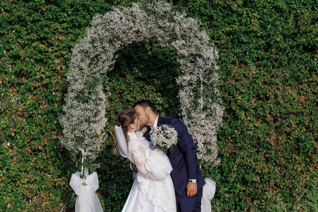 Bride and groom kissing under a floral wedding arch during a garden ceremony in Alessandria, Piedmont, Italy