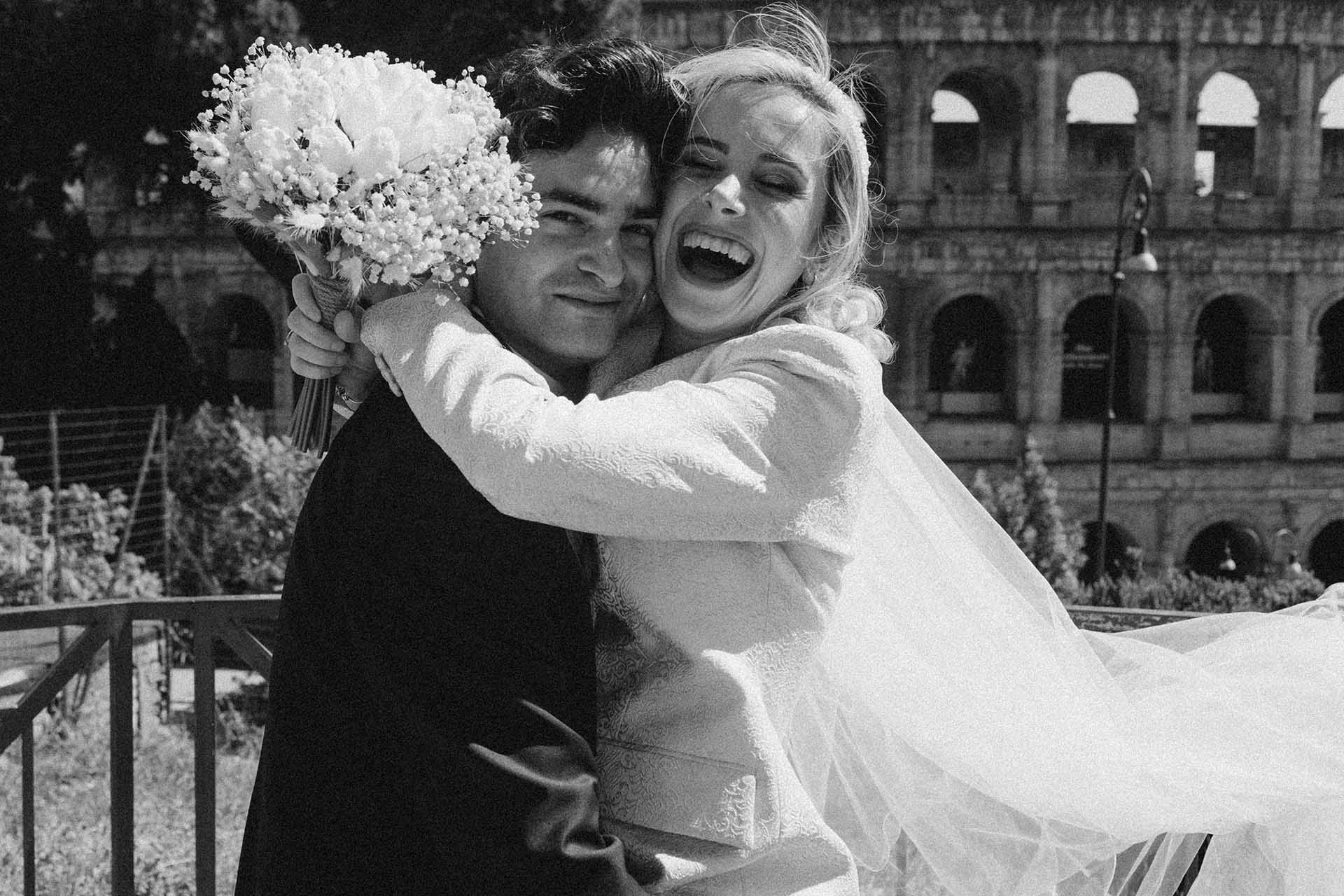 Black and white portrait of joyful bride embracing groom in Rome with historic Roman architecture in the background