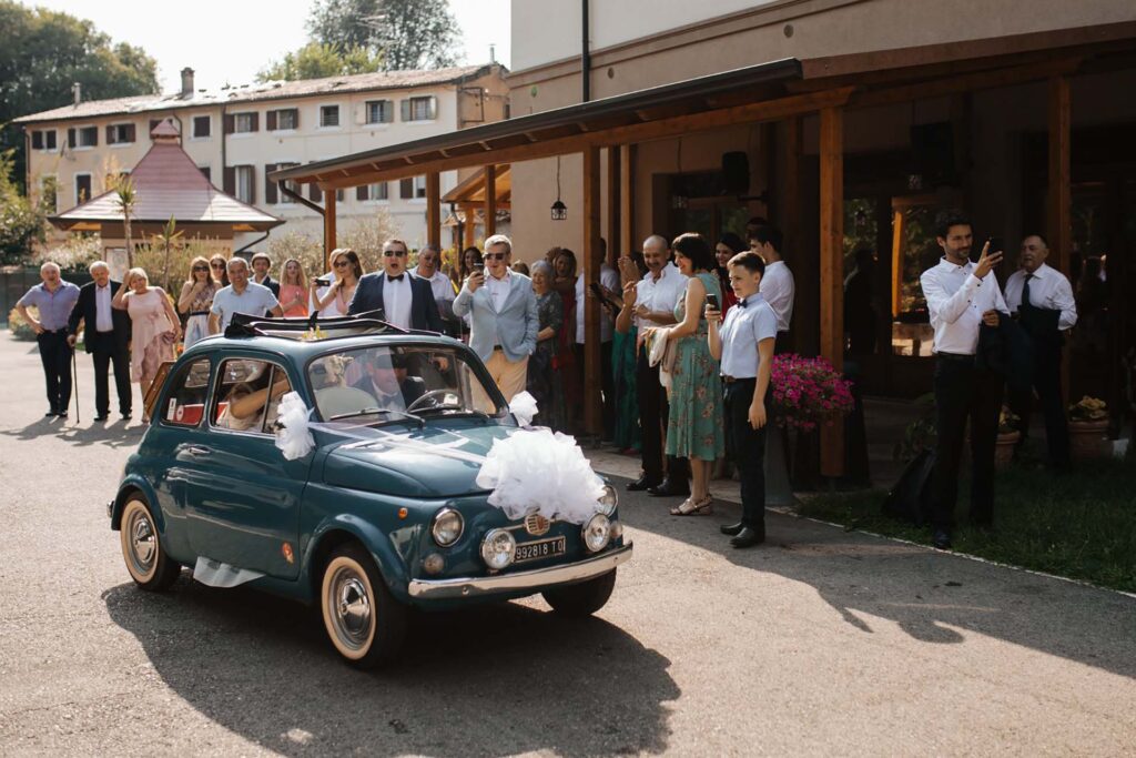 Bride and groom departing in a vintage Fiat 500 during a countryside wedding celebration in Italy surrounded by guests