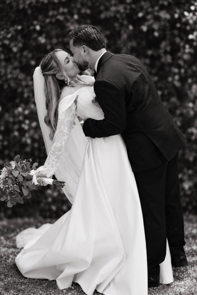 Black-and-white portrait of the bride and groom kissing outdoors after their wedding ceremony