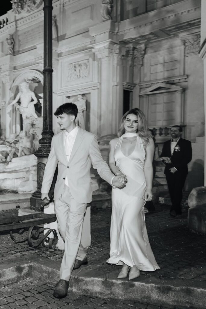 Elegantly dressed wedding guests walking beside the illuminated fountain during an evening reception in Rome