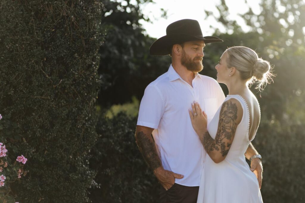 Romantic couple embracing in warm golden-hour sunlight, featuring a bride in a white open-back dress with floral tattoos and a bearded man wearing a black wide-brimmed hat