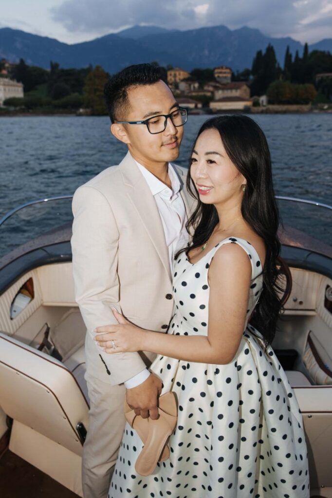 Woman in a white polka dot dress posing aboard a classic wooden motorboat on Lake Como, with mountains and Italian lakeside villas behind her