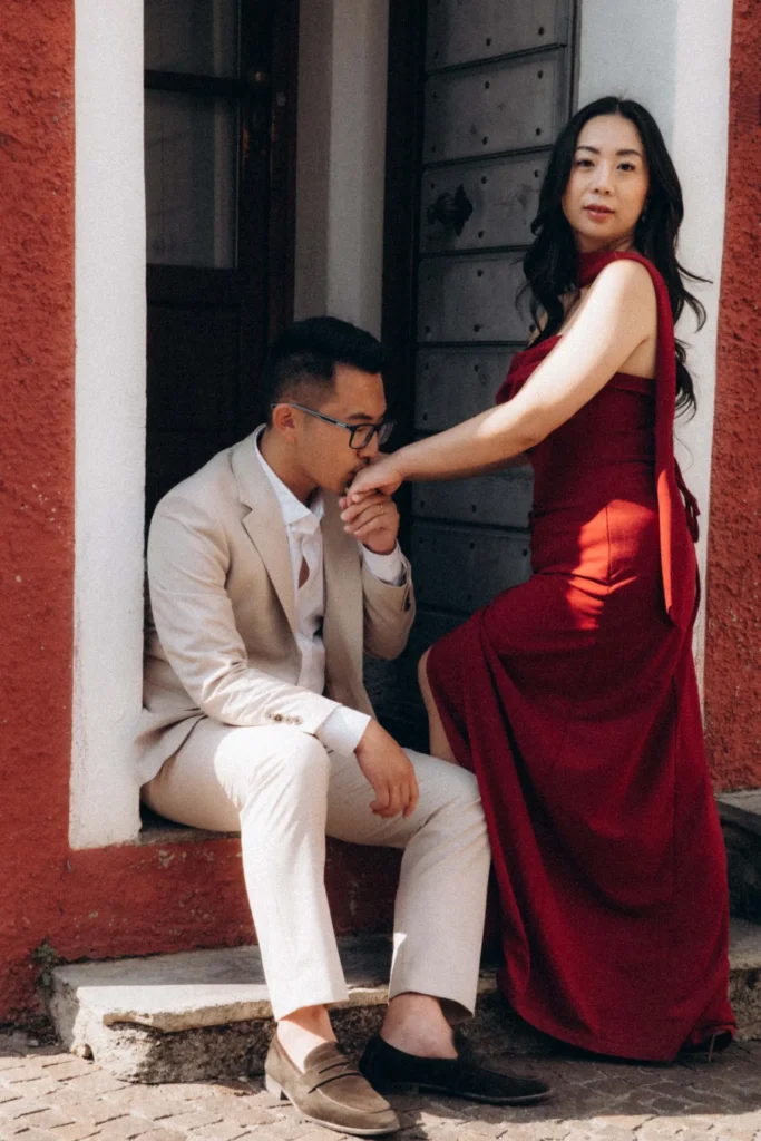 Man in a light suit kissing his partner's hand as she wears a red gown beside a stone doorway