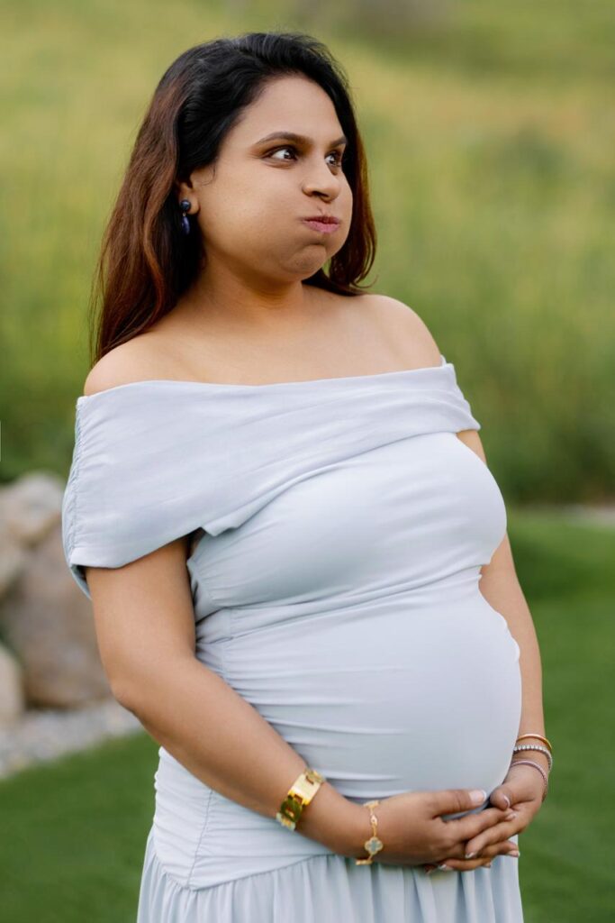 Expectant mother in an elegant white off-the-shoulder maternity dress, gently holding her baby bump and making a playful expression during an outdoor photoshoot