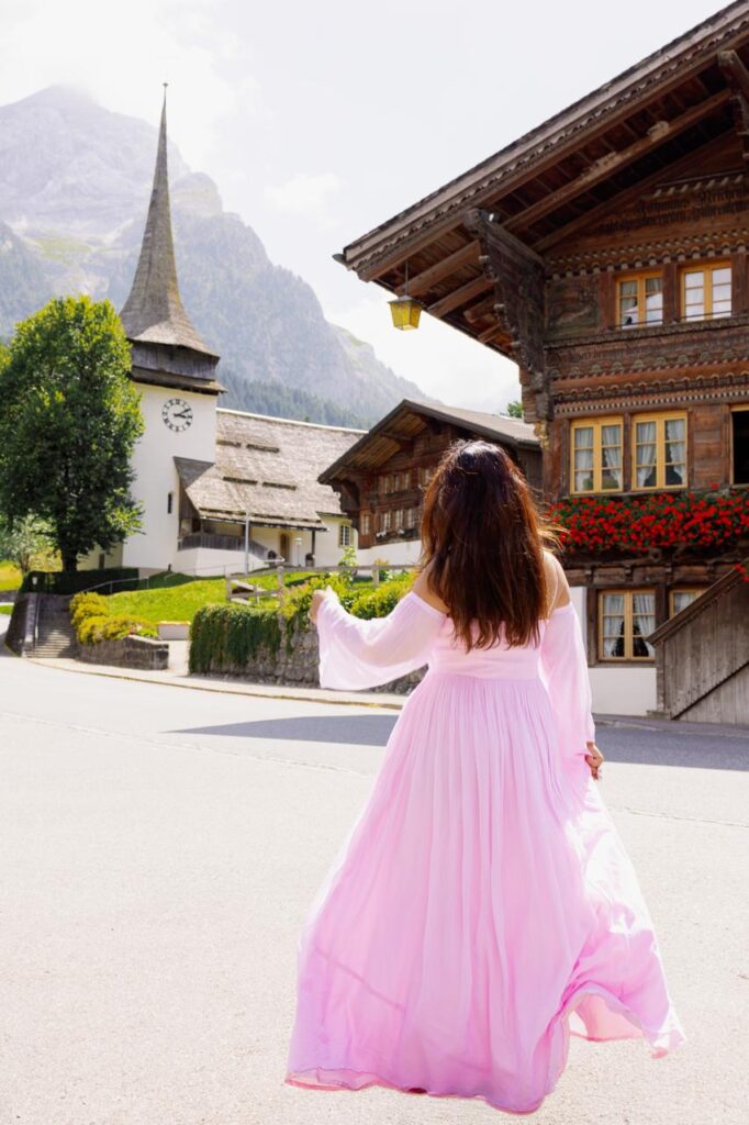 Woman in a flowing pink dress walking through a picturesque Swiss Alpine village, surrounded by traditional wooden chalets, a historic church and dramatic mountain scenery.